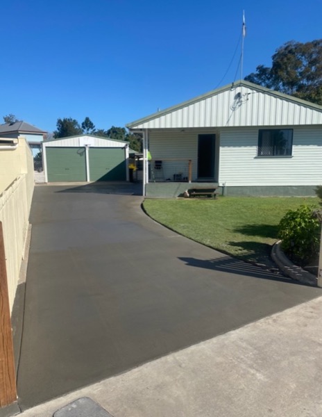 A plain grey concrete driveway running past a shed to a weatherboard house