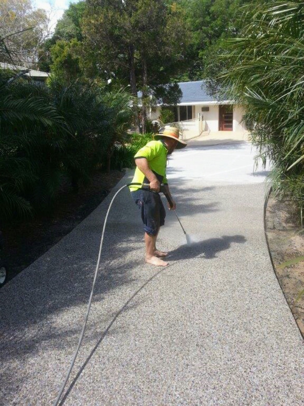 A worker in a wide-brim hat washing down a concrete driveway with a hose